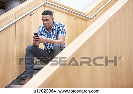 Picture - Young businessman sitting on office stairway texting on smartphone. Fotosearch