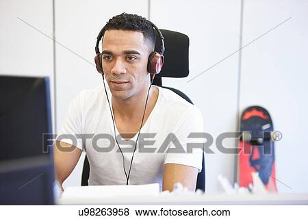 Stock Photo - Young businessman using computer whilst listening to headphones in office. Fotosearch