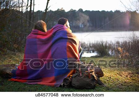 Stock Photo - Young couple at lakeside wrapped in blanket at sunset. Fotosearch