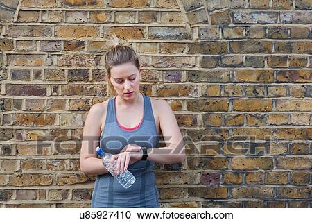 Stock Image - Young female runner leaning against brick wall checking stop watch. Fotosearch