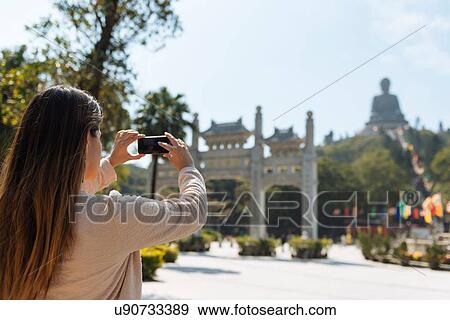 Stock Photo - "Young female tourist taking smartphone photographs of Tian Tan Buddha, Po Lin Monastery, Lantau Island, Hong Kong, China". Fotosearch