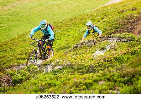 Stock Image - Young male and female mountain bikers cycling on hillside track. Fotosearch