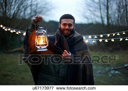 Stock Photo - Young male camper holding up lantern whilst wrapped in blanket in woods. Fotosearch