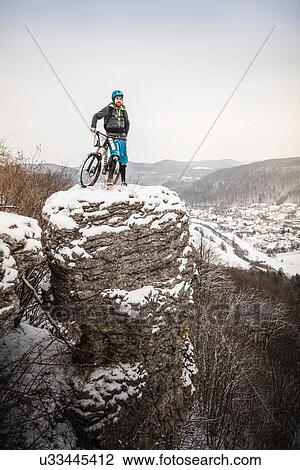 Stock Image - Young male mountain biker on top of snow covered rock formation. Fotosearch