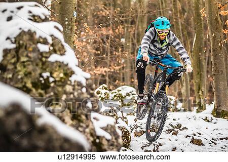 Stock Photography - Young male mountain biker speeding on snow covered rocks in forest. Fotosearch