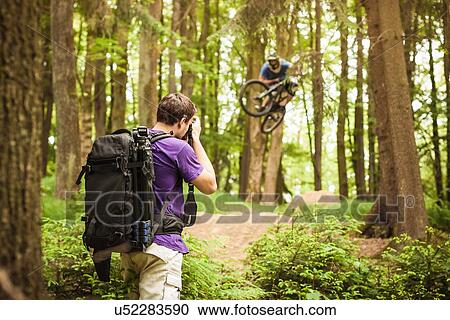 Stock Image - Young male photographer photographing mountain biker in forest. Fotosearch