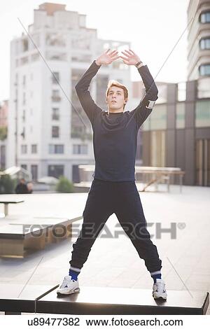 Stock Image - Young male runner stretching on bench in city square. Fotosearch