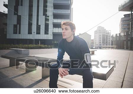 Stock Image - Young male runner taking a break on bench in city square. Fotosearch
