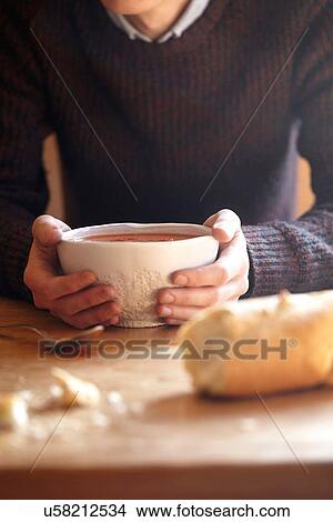 Picture - Young man at kitchen table with hands holding soup bowl. Fotosearch
