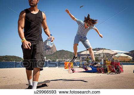 Stock Image - "Young man doing jump on skateboard, while friend looks away". Fotosearch