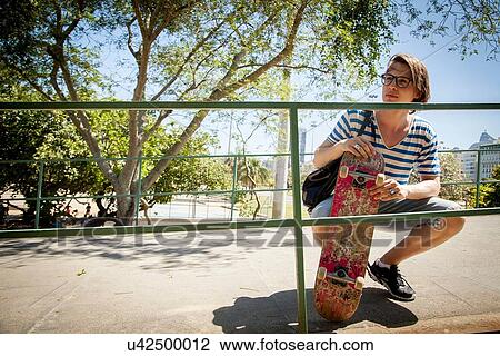 "Young man holding skateboard, crouching beside railings" View Large Photo Image Stock Image - "Young man holding skateboard, crouching beside railings". Fotosearch