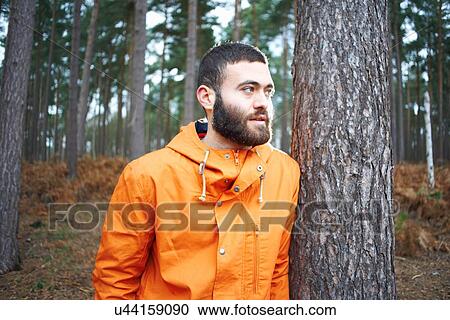 Stock Image - Young man leaning against tree looking out from forest. Fotosearch