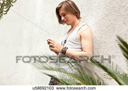 Stock Image - Young man leaning against wall reading text messages on smartphone. Fotosearch