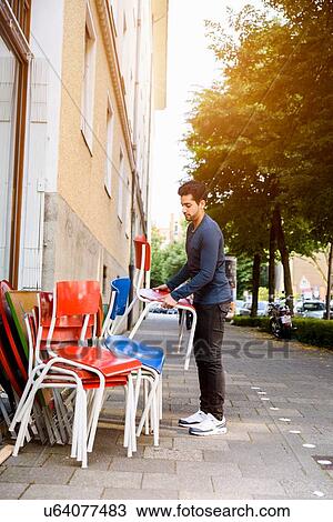 Stock Image - "Young man, putting chairs out, outside building". Fotosearch