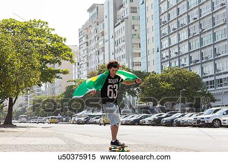 Stock Photography - "Young man skateboarding holding Brazilian flag, Copacabana, Rio De Janeiro, Brazil". Fotosearch