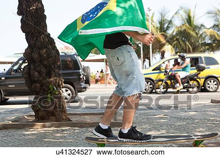 Stock Photo - "Young man skateboarding wrapped in Brazilian flag, Copacabana, Rio De Janeiro, Brazil". Fotosearch