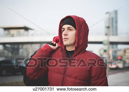 Stock Image - Young man wearing red hooded anorak on city street. Fotosearch