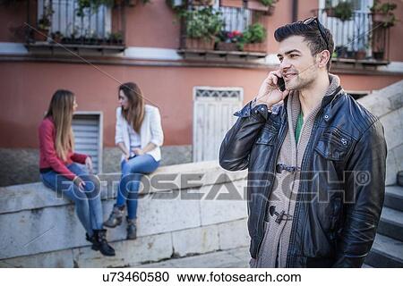 Stock Image - "Young man with friends talking on smartphone, Cagliari, Sardinia, Italy". Fotosearch
