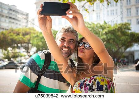 Stock Image - "Young woman and mature man taking smartphone selfie, Copacabana, Rio De Janeiro, Brazil". Fotosearch