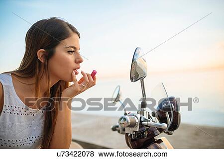 Stock Photo - "Young woman applying lipstick in motorcycle mirror, Manila, Philippines". Fotosearch