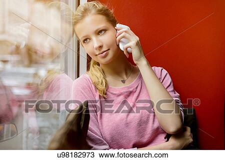 Stock Image - Young woman gazing out of cafe window whilst chatting on smartphone. Fotosearch
