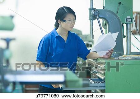Stock Image - Young woman holding papers in industrial workshop. Fotosearch