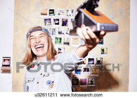 Stock Image - Young woman in front of photo wall taking instant selfie on retro camera. Fotosearch