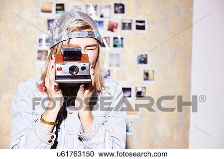 Stock Image - Young woman in front of photo wall taking photograph on retro camera. Fotosearch