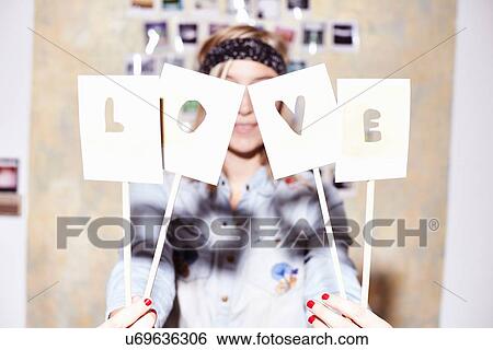 Young woman in front of photo wall hiding behind love sign View Large Photo Image Stock Photograph - Young woman in front of photo wall hiding behind love sign. Fotosearch