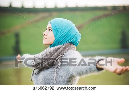 Stock Image - Young woman in park with arms open wearing turquoise hijab. Fotosearch