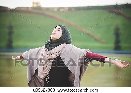 Stock Image - Young woman in park with arms open wearing hijab. Fotosearch