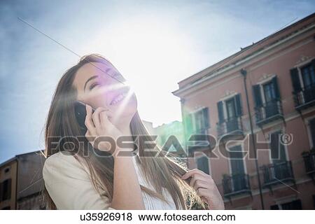 Stock Photo - "Young woman in sunlit street talking on smartphone, Cagliari, Sardinia, Italy". Fotosearch