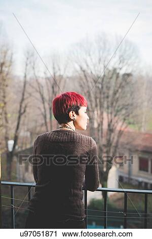 Stock Image - "Young woman leaning against balcony railings, looking at view". Fotosearch
