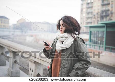 Stock Image - Young woman leaning against wall reading smartphone texts. Fotosearch