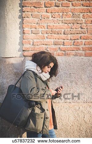 Stock Image - Young woman leaning against wall reading smartphone texts. Fotosearch