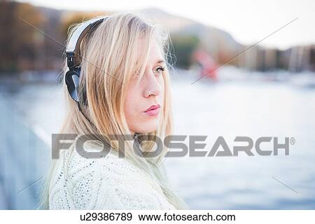 Stock Photo - "Young woman looking away whilst listening to headphones on lakeside, Como, Italy". Fotosearch