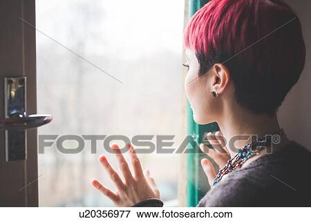 Stock Photo - "Young woman looking out of window, hands touching window, rear view". Fotosearch