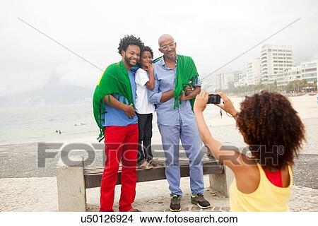 Picture - "Young woman photographing family wrapped in Brazilian flag on Ipanema beach, Rio De Janeiro, Brazil". Fotosearch