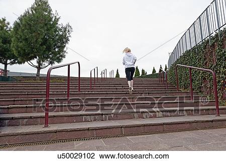 Stock Image - "Young woman running up steps, rear view". Fotosearch