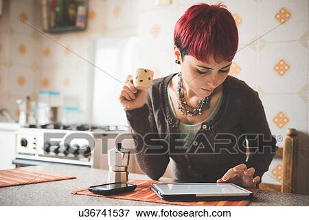Stock Photo - "Young woman sitting at table drinking coffee, using digital tablet". Fotosearch