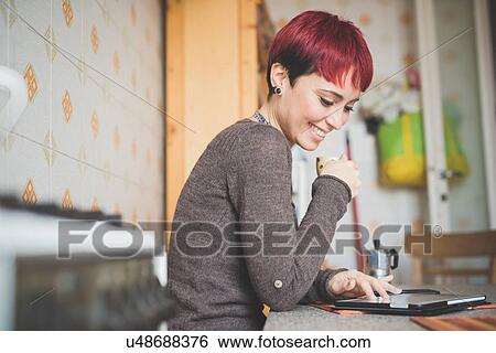 Stock Photograph - "Young woman sitting at table drinking coffee, looking at digital tablet". Fotosearch