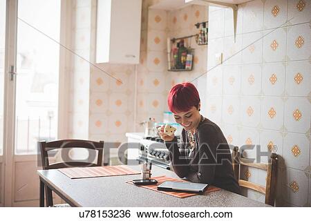 Stock Photograph - "Young woman sitting at table drinking coffee, looking at digital tablet". Fotosearch