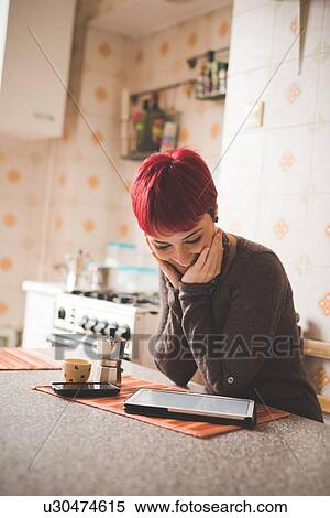 Stock Photography - Young woman sitting at table looking at digital tablet. Fotosearch
