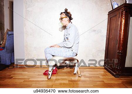 Stock Image - Young woman sitting on hassock wearing sunglasses and blowing bubblegum. Fotosearch