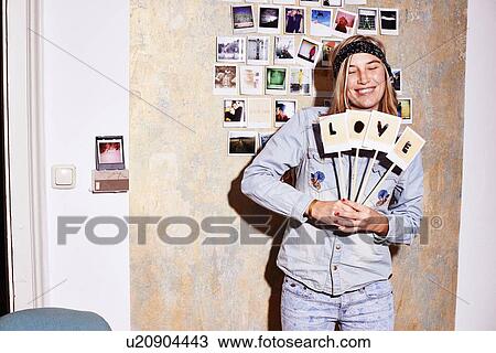 Stock Image - Young woman standing in front of photograph wall holding up love sign. Fotosearch