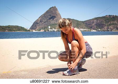 Stock Photo - "Young woman tying shoelace on beach, Rio de Janeiro, Brazil". Fotosearch