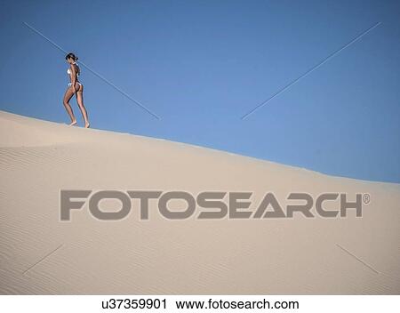 Stock Image - Young woman walking on sand dune against clear blue sky. Fotosearch
