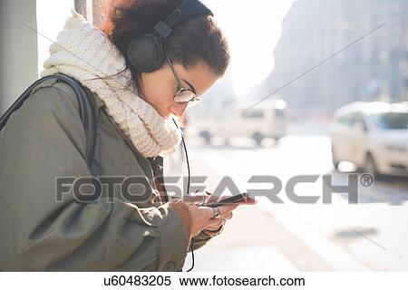 Stock Photography - Young woman wearing headphones choosing smartphone music on street. Fotosearch