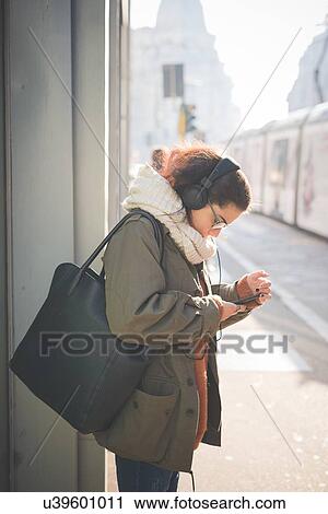 Young woman wearing headphones reading smartphone texts on street View Large Photo Image Stock Image - Young woman wearing headphones reading smartphone texts on street. Fotosearch