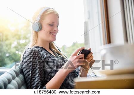 Stock Image - "Young woman, wearing headphones, sitting in cafe, holding smartphone". Fotosearch